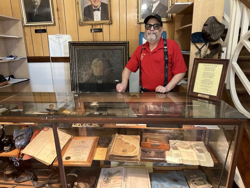 Historical Curator Bro. Lee Hoffnung behind the museum's glass display case, beside a portrait of King Lodge's founder
